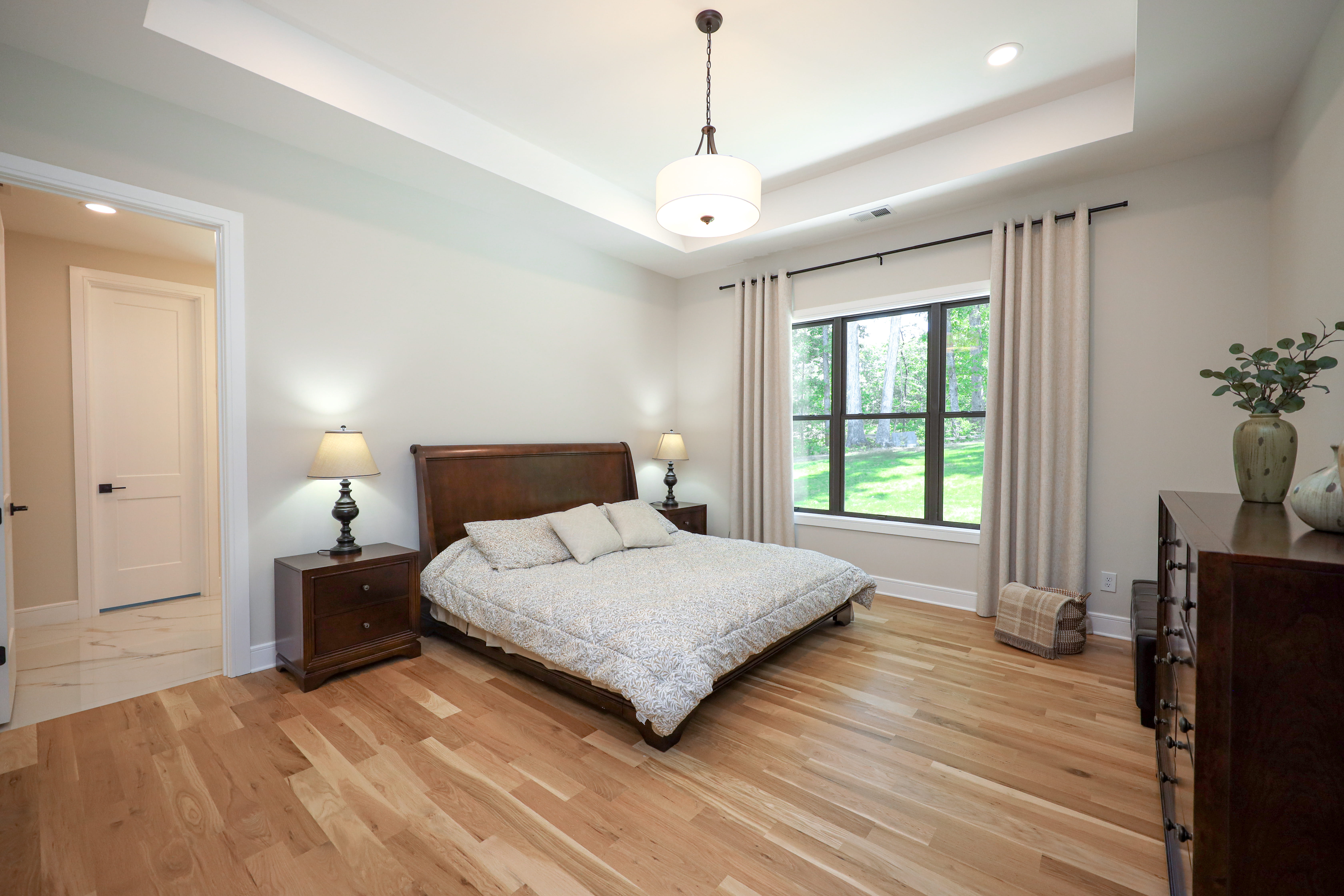 Primary bedroom with a tray ceiling, hardwood floors, neutral decor, and black-framed windows overlooking a wooded yard.