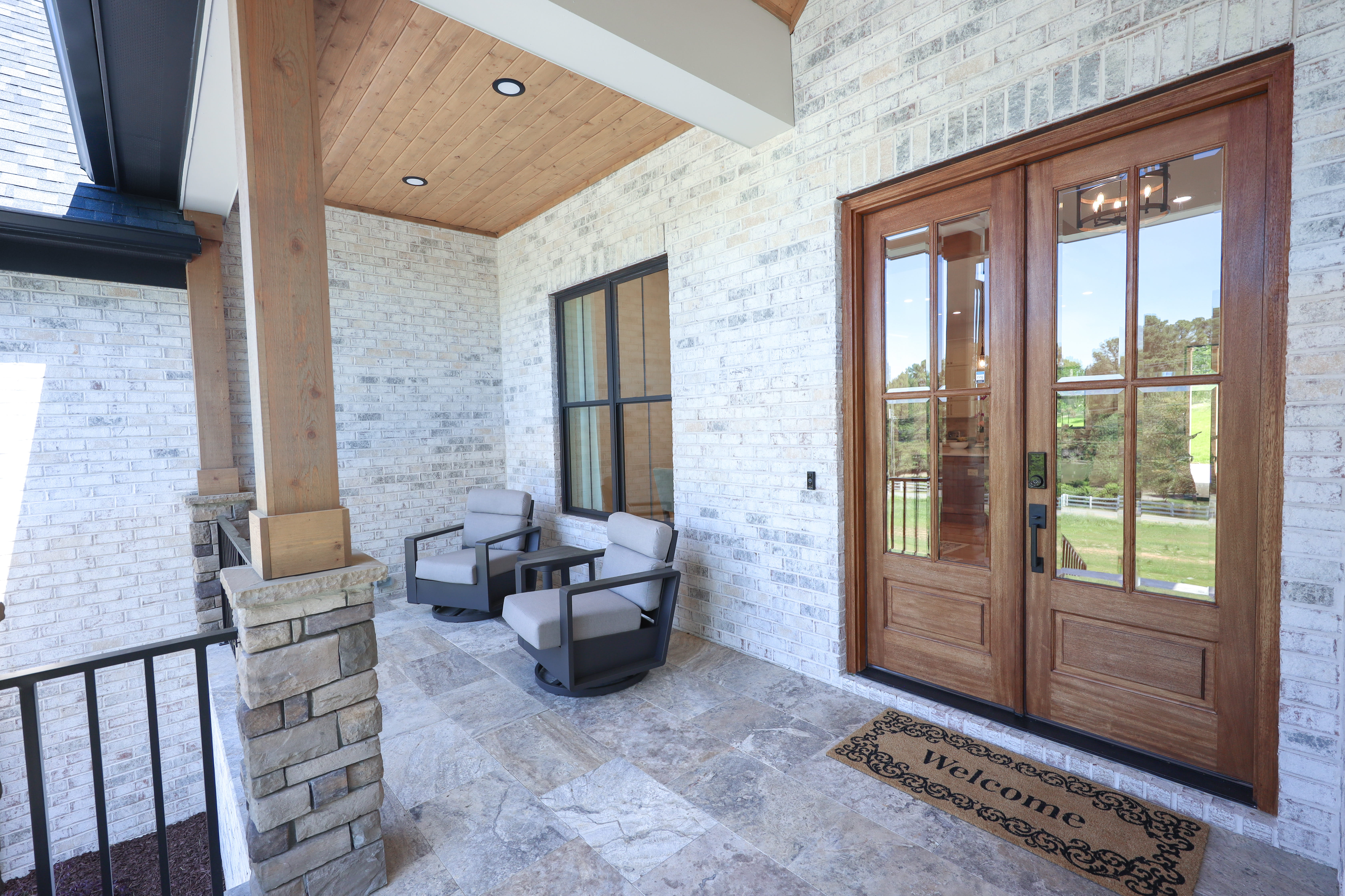 Covered front porch with timber posts, white brick, stone flooring, seating area, and glass-paneled wood double doors.