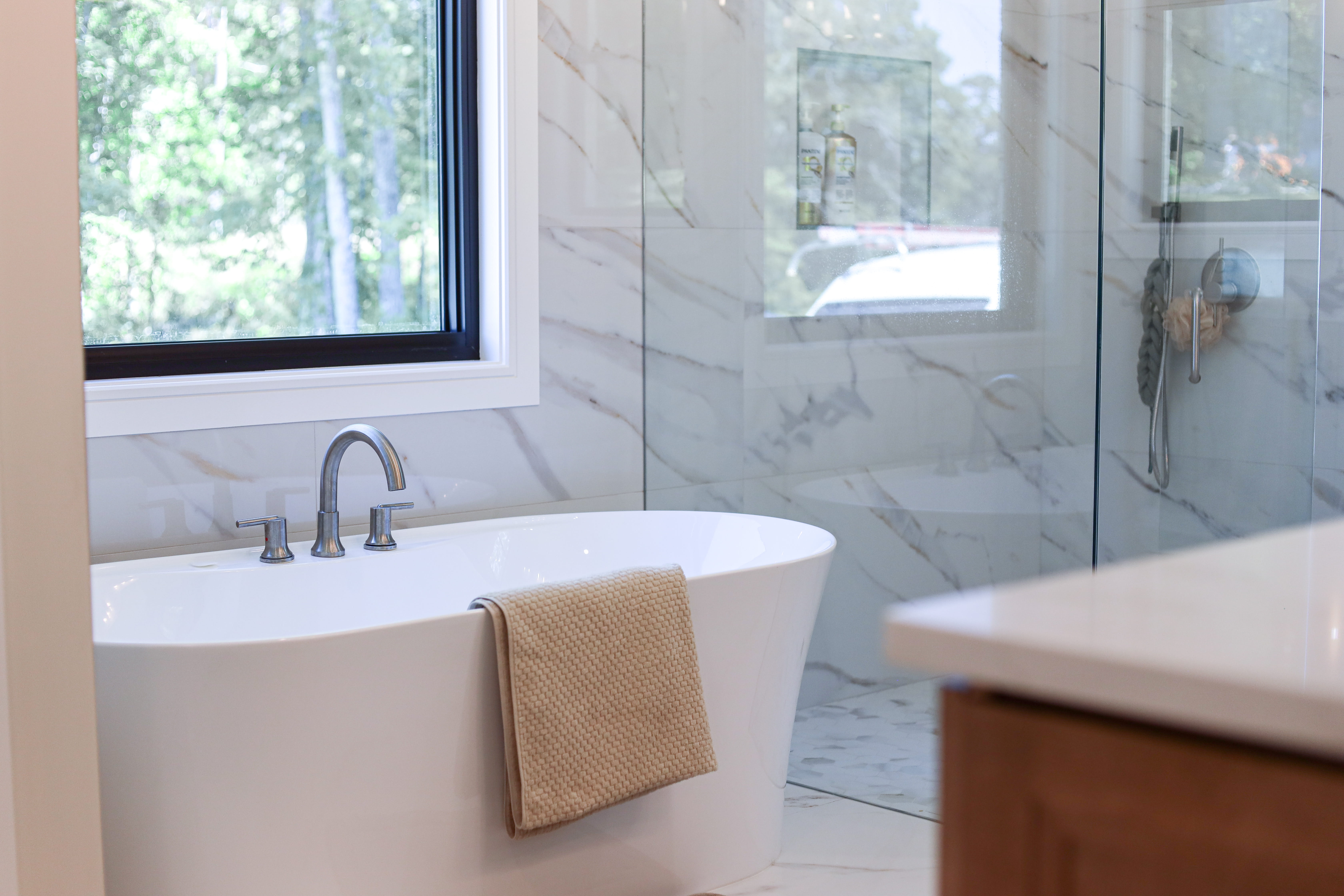 Primary bathroom with a freestanding soaking tub, marble-look tile, black-framed window, and glass-enclosed shower.